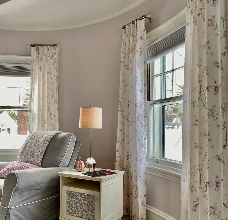 Living room with floral curtains, grey sofa, white side table, and lamp. Windows with outside view.