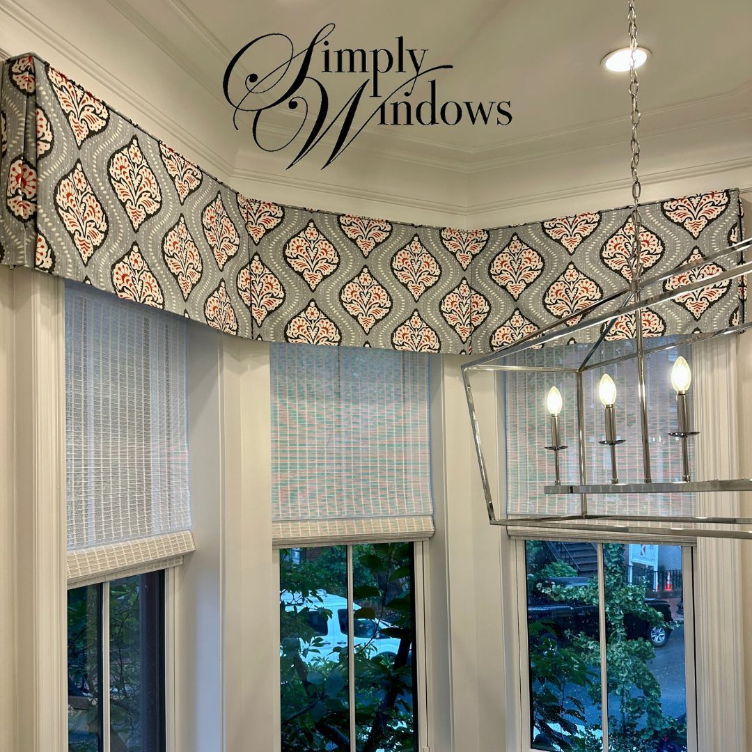 A bay window featuring a patterned grey and coral fabric valance over white woven Roman shades and a metal chandelier.
