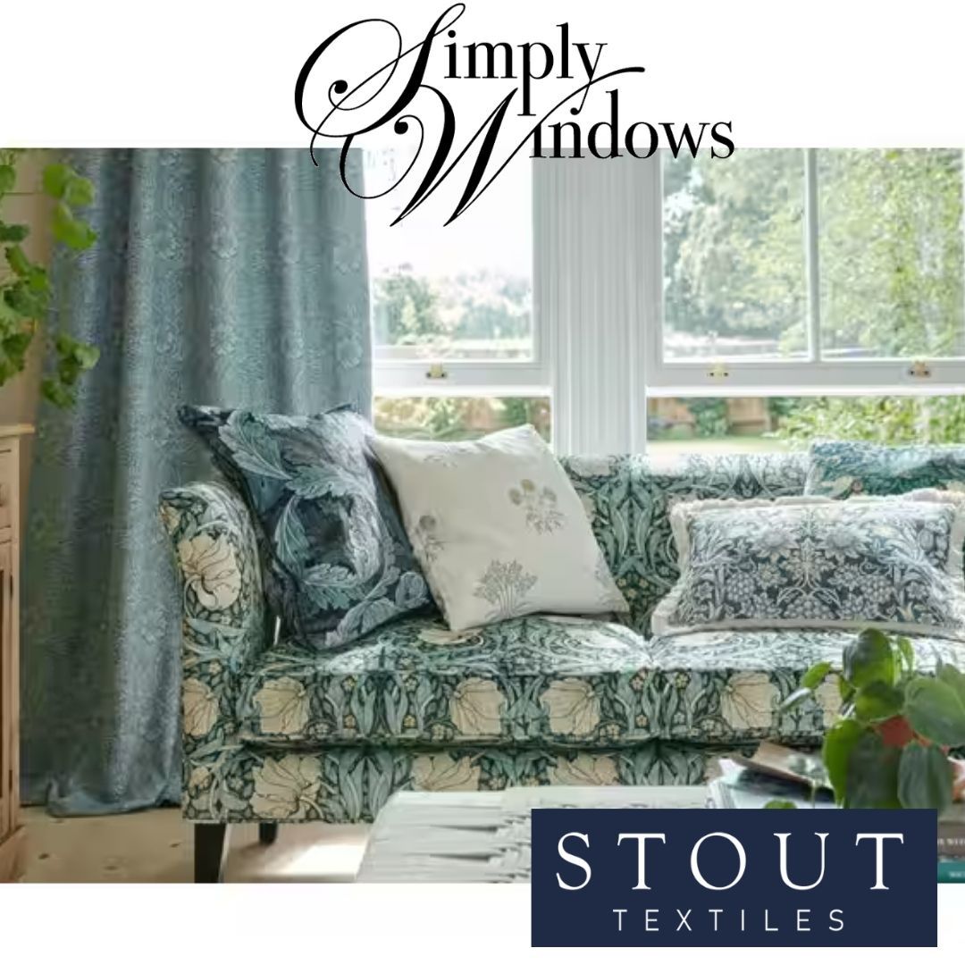 A patterned blue sofa with decorative pillows stands next to a matching curtain in a room with large windows.