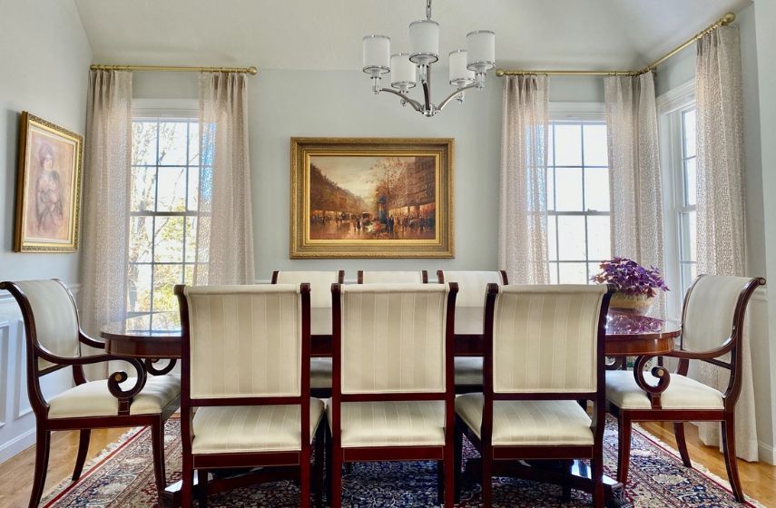 A dining room with several windows has gold decorative rods and beige drapes.  