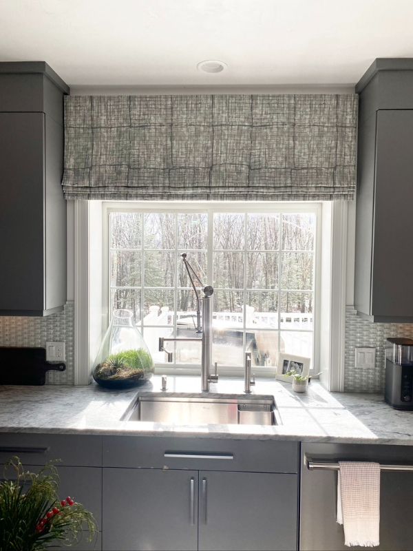 A grey kitchen has patterned Roman shades above the window over the sink. 