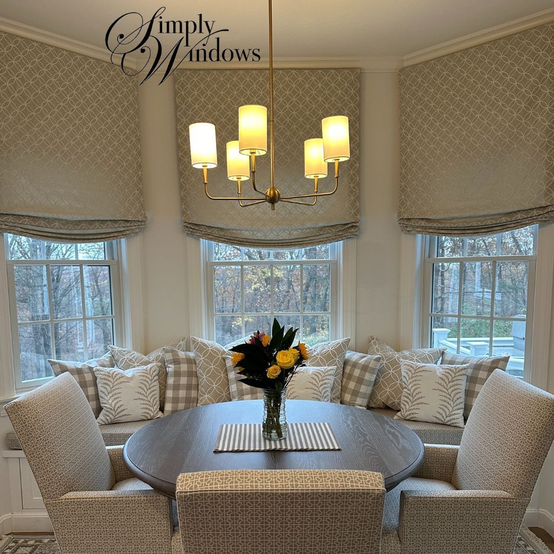 A dining nook with a round table, chairs, a built-in bench with patterned pillows, and roman shades under a chandelier.