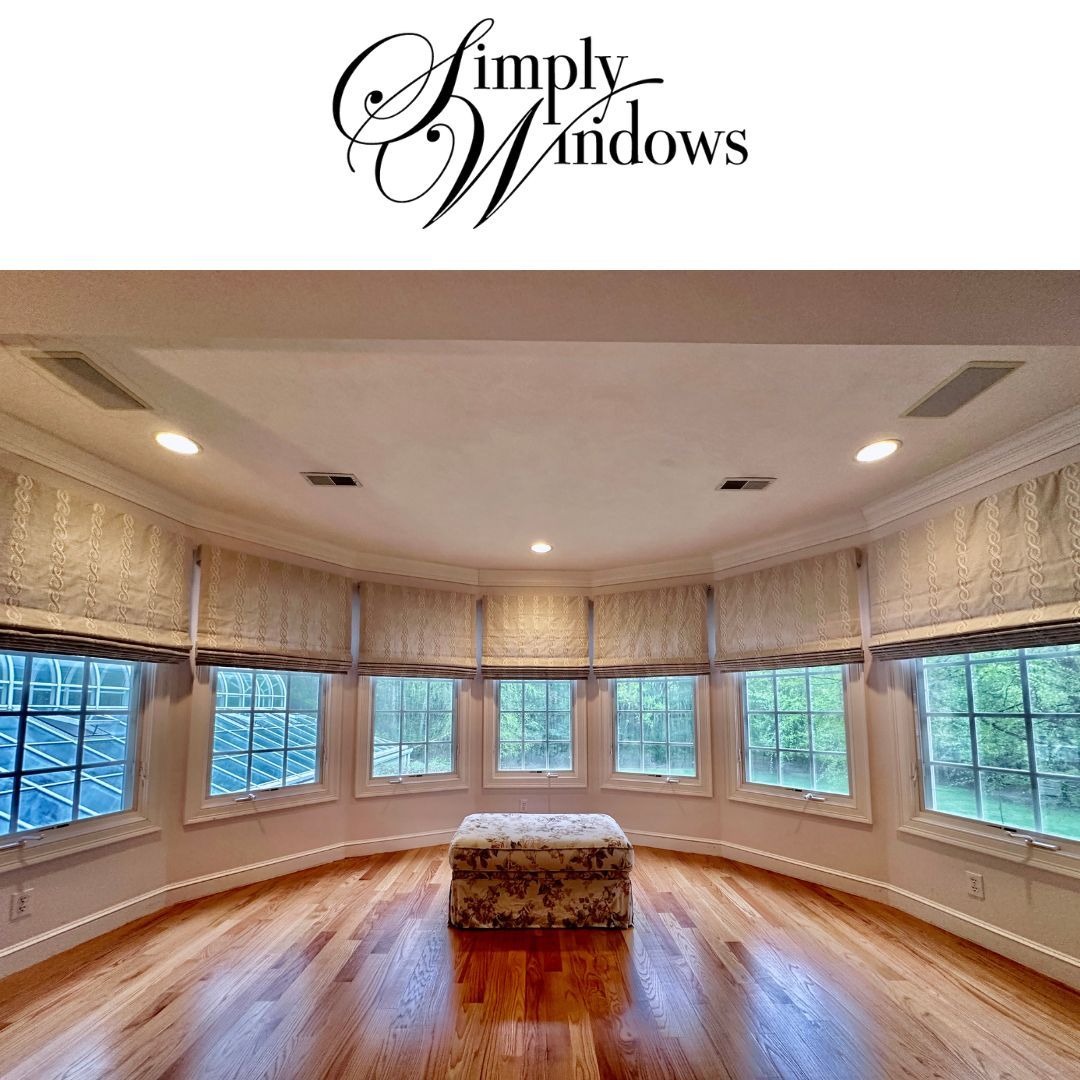 A curved bay window room with hardwood floors, light-colored Roman shades, and a central floral ottoman.
