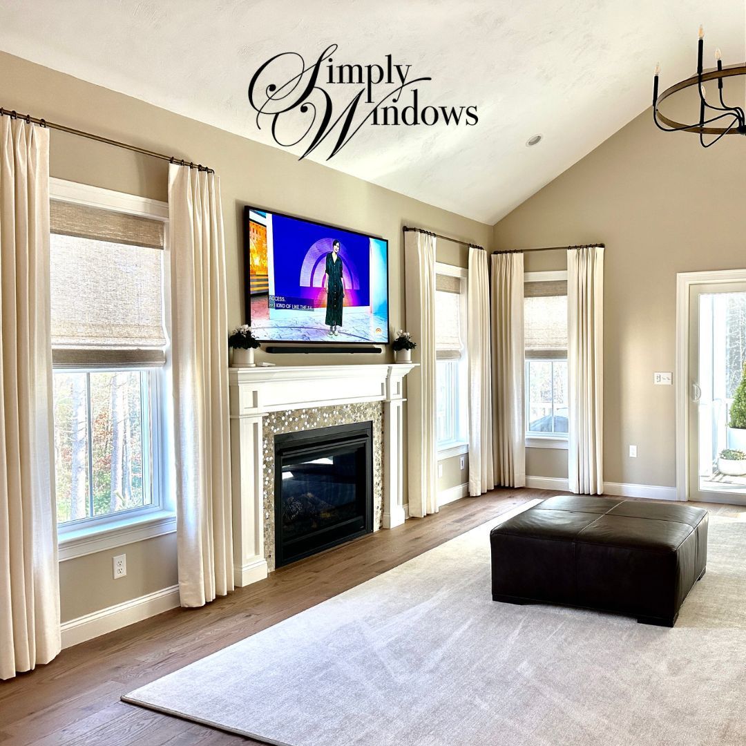 Bright living room with fireplace, TV, layered window treatments on large windows, and dark ottoman on a light rug