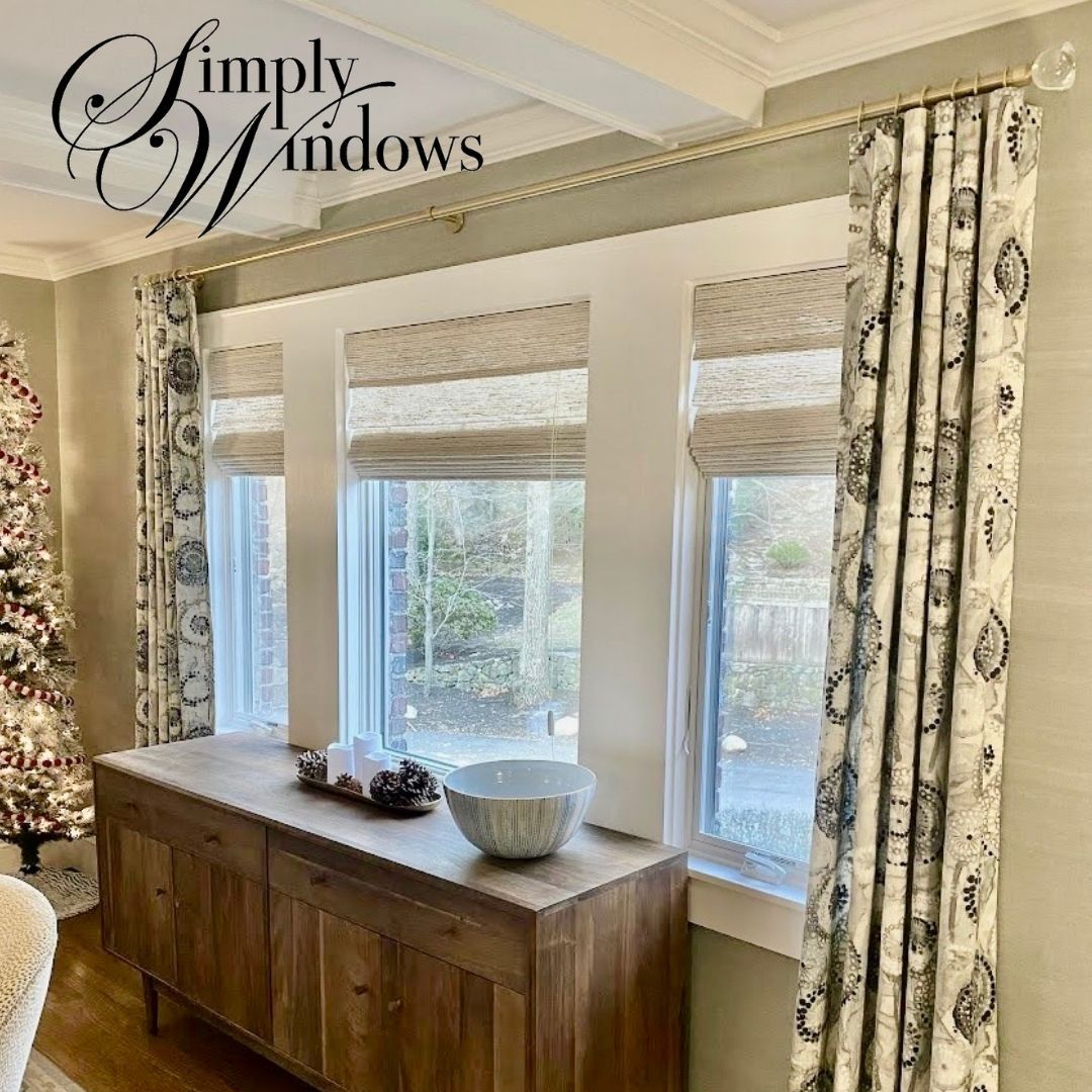 A wooden sideboard sits below three windows with natural woven shades and patterned, floor-length drapery on a gold rod.