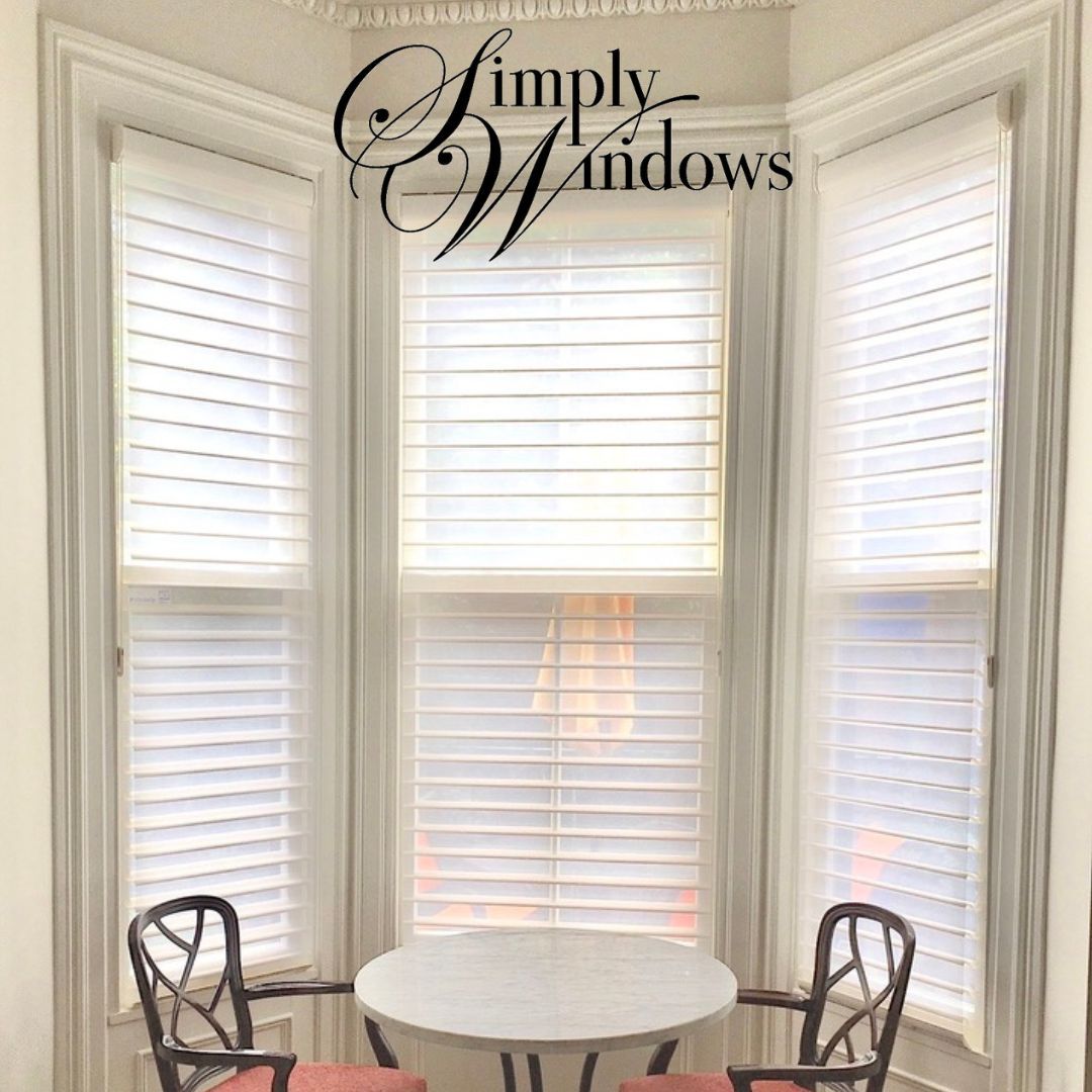 A bay window with white plantation shutters, featuring a small round table and two decorative chairs in front.