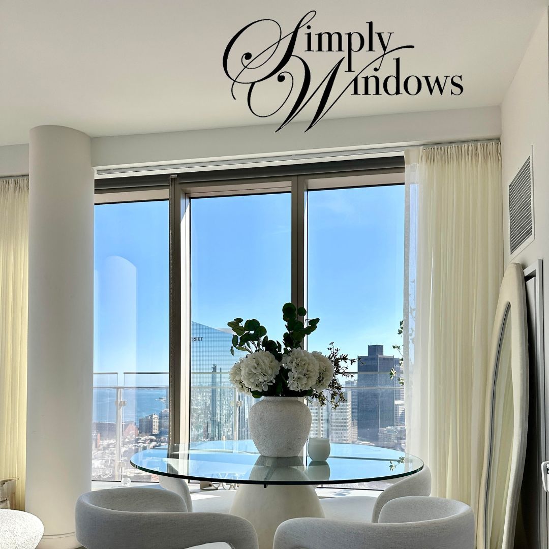 A dining area with a glass table, white chairs, and a flower vase, set against large windows overlooking a city skyline.