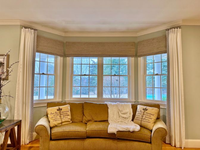 A living room has a brown counch in front of a bay window with drapes and woven wood shades. 