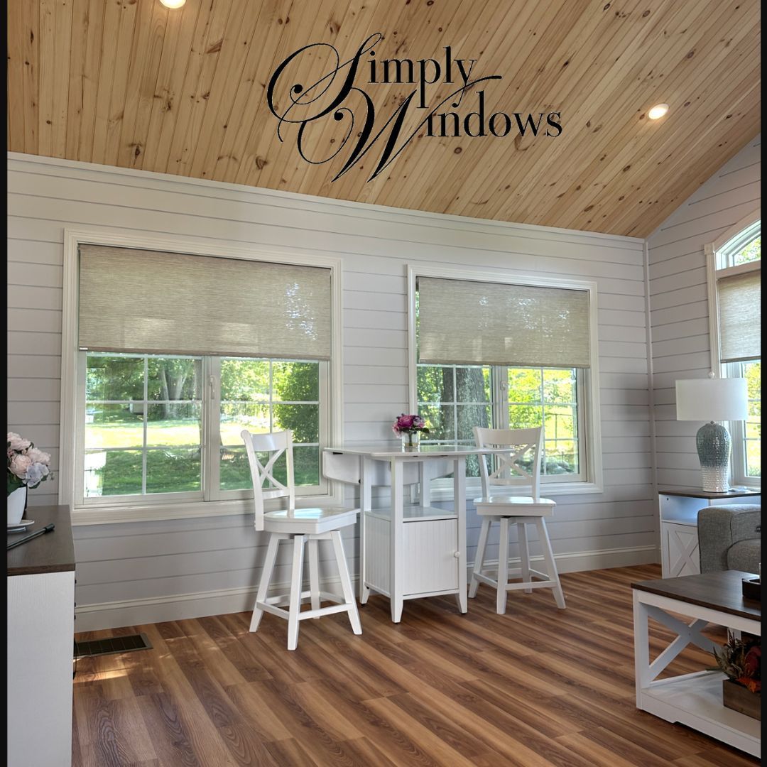 A bright sunroom with white shiplap walls, a wood plank ceiling, two windows with roller shades, and a white bistro set.