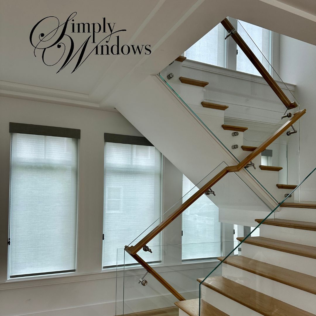 Interior with white walls, staircase with wooden and glass railing, windows with gray shades.