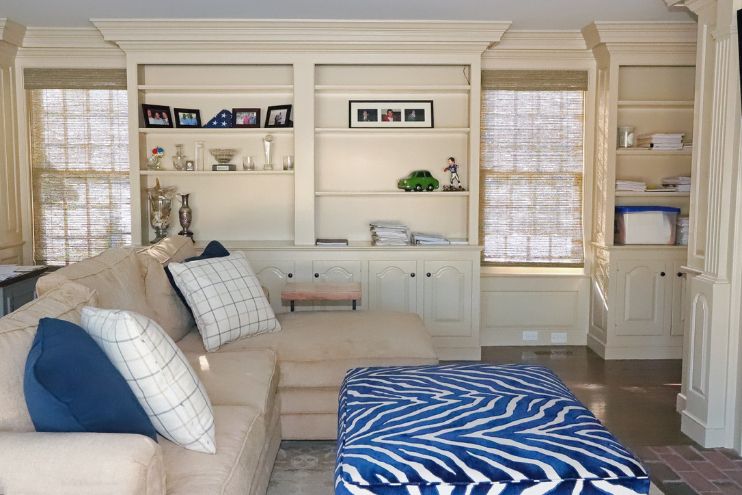 A den area with beige furniture and blue accent pillows has windows with woven wood shades. 