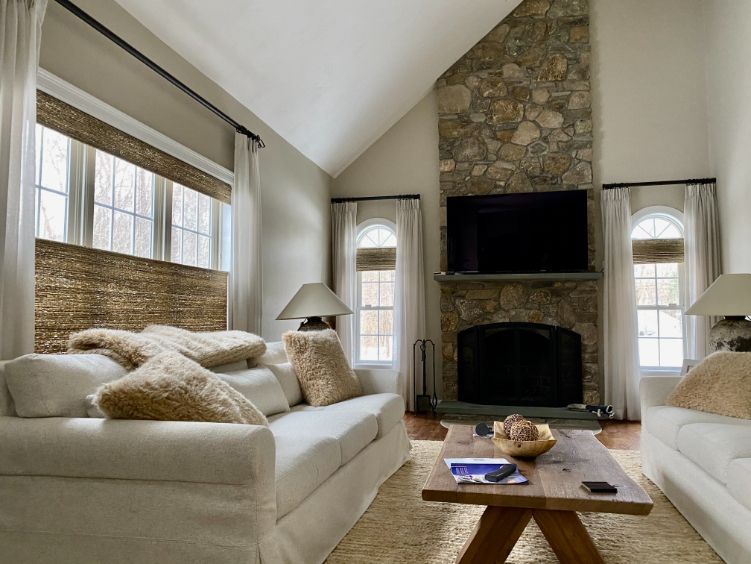 A large living room with a stone fireplace has windows with bamboo roller shades. 