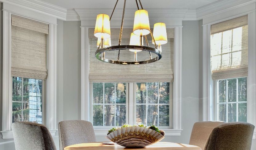 A dining room has three windows behind it with woven wood bamboo roman shades. 