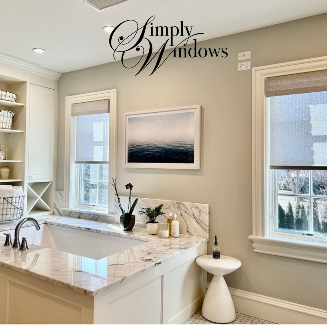 A marble-topped bathtub in a neutral-toned bathroom with two windows featuring light-filtering shades and framed wall art.