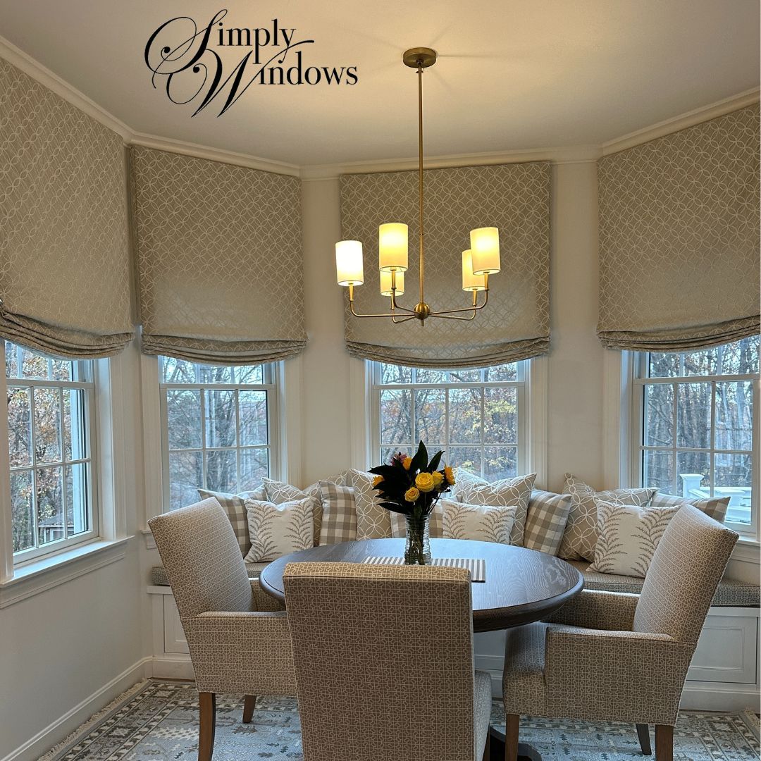 A dining nook with a round table, chairs, and cushioned bench seating under three windows with patterned Roman shades.