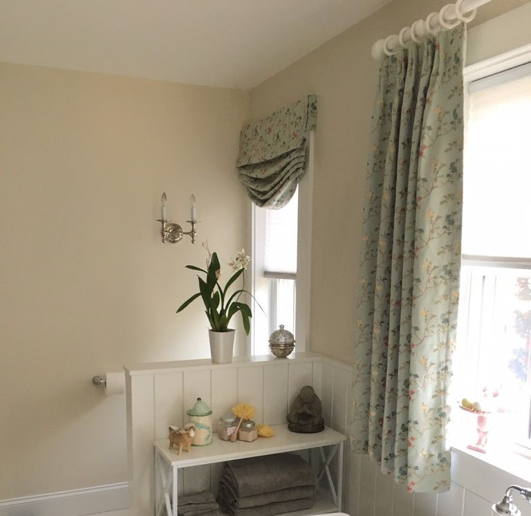 A corner of a room features beige walls, floral curtains, a Roman shade, and a small white shelf holding towels and decor.
