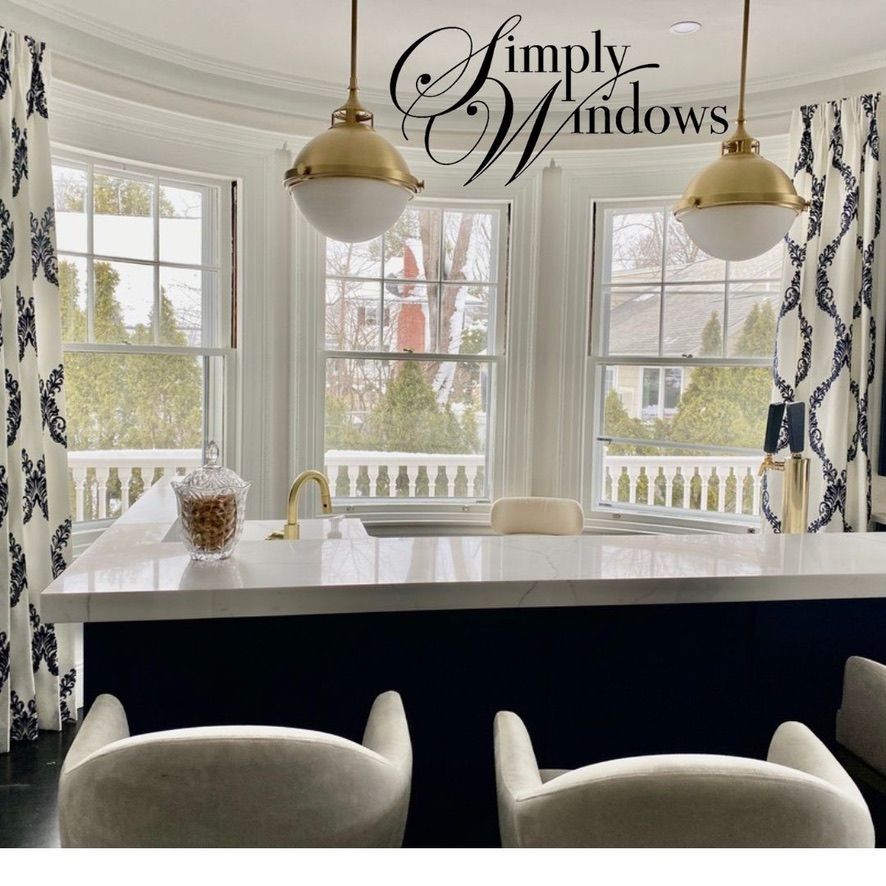 A bar area with a white countertop, two stools, brass lights, and floor-length patterned curtains in front of bay windows.