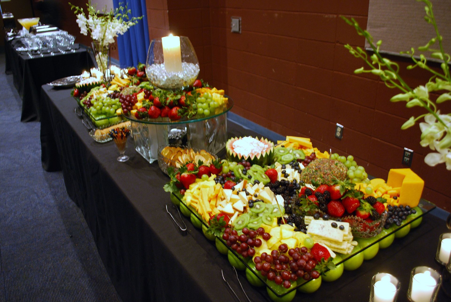 Buffet table with trays of fruit, cheeses, and hors d'oeuvres along a brick wall