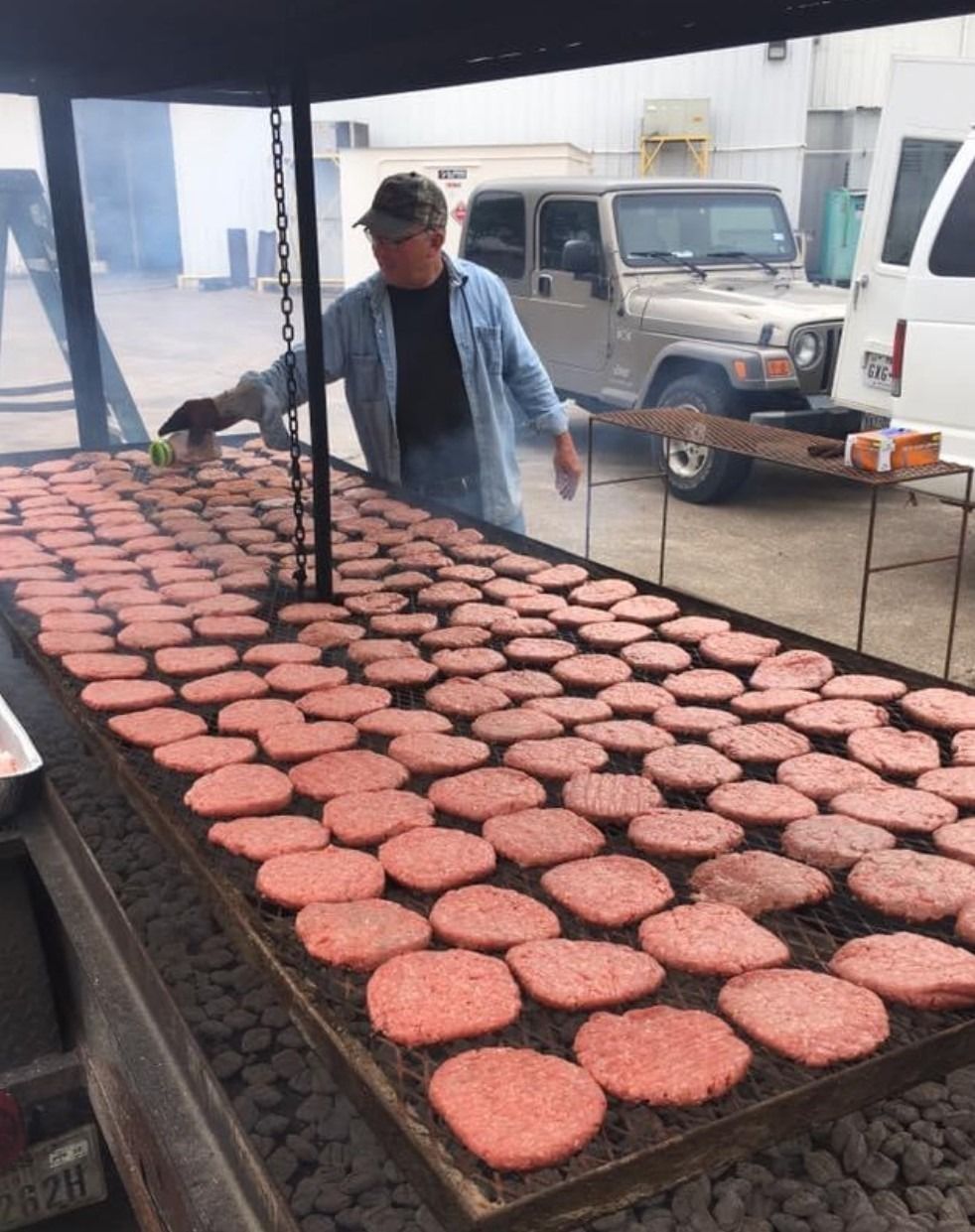 Person grilling rows of hamburger patties on a large outdoor barbecue with a truck in the background