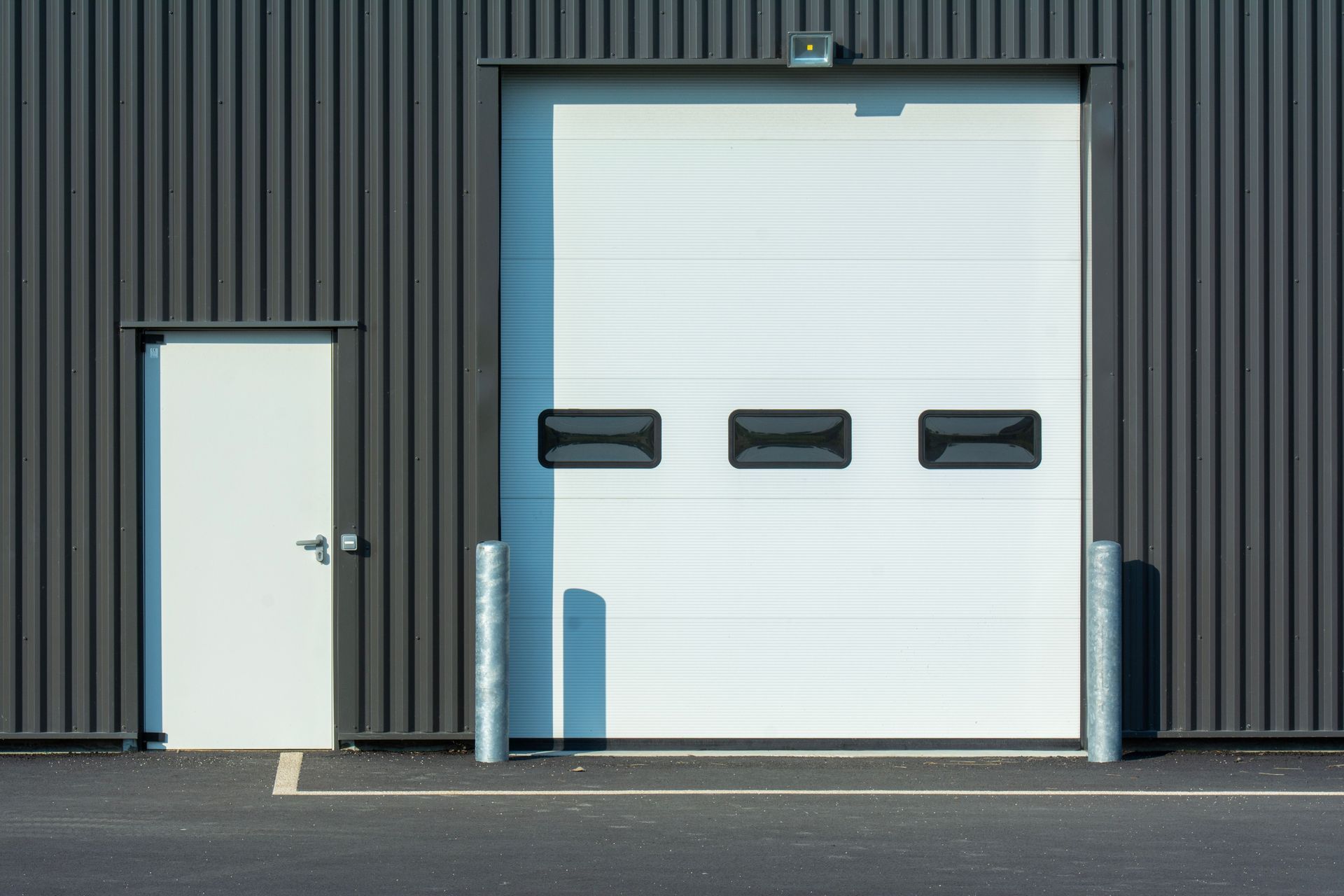 Gray industrial building with a white side door and large garage door with small windows.