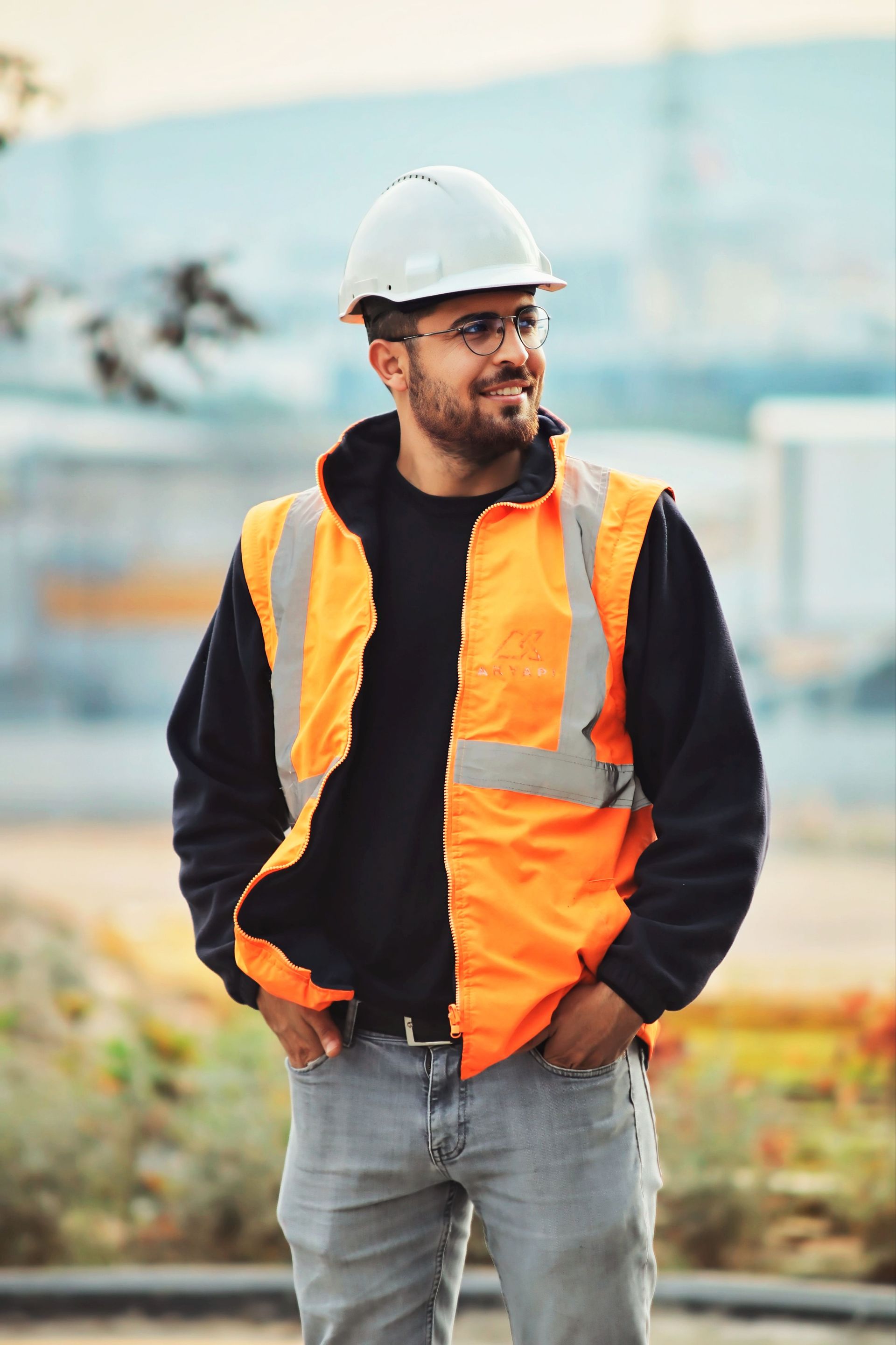 Worker in a white hard hat and orange safety vest standing outdoors with hands in pockets.