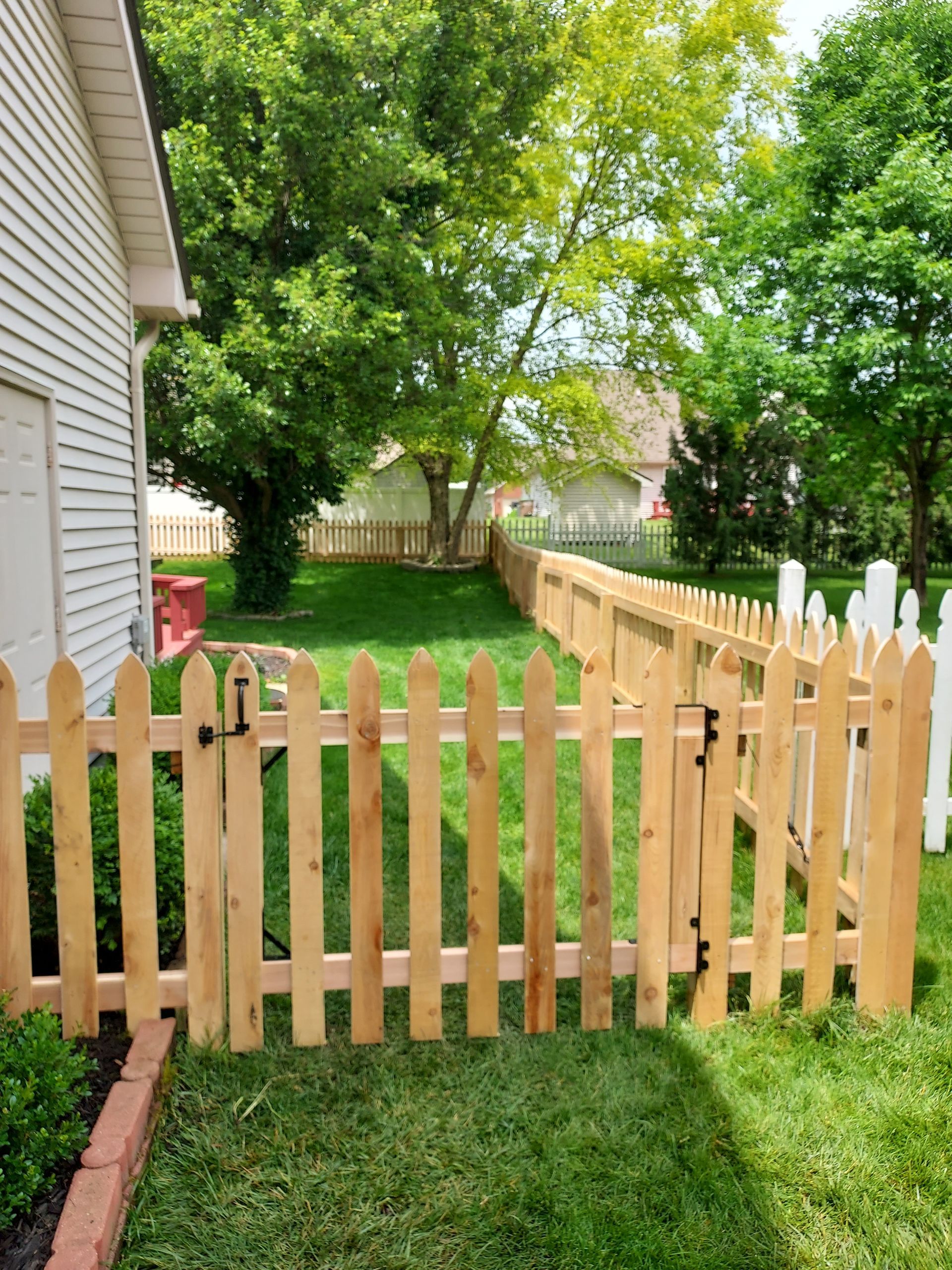 Wooden picket fence, partially enclosing a grassy backyard with trees and neighboring houses in the background.