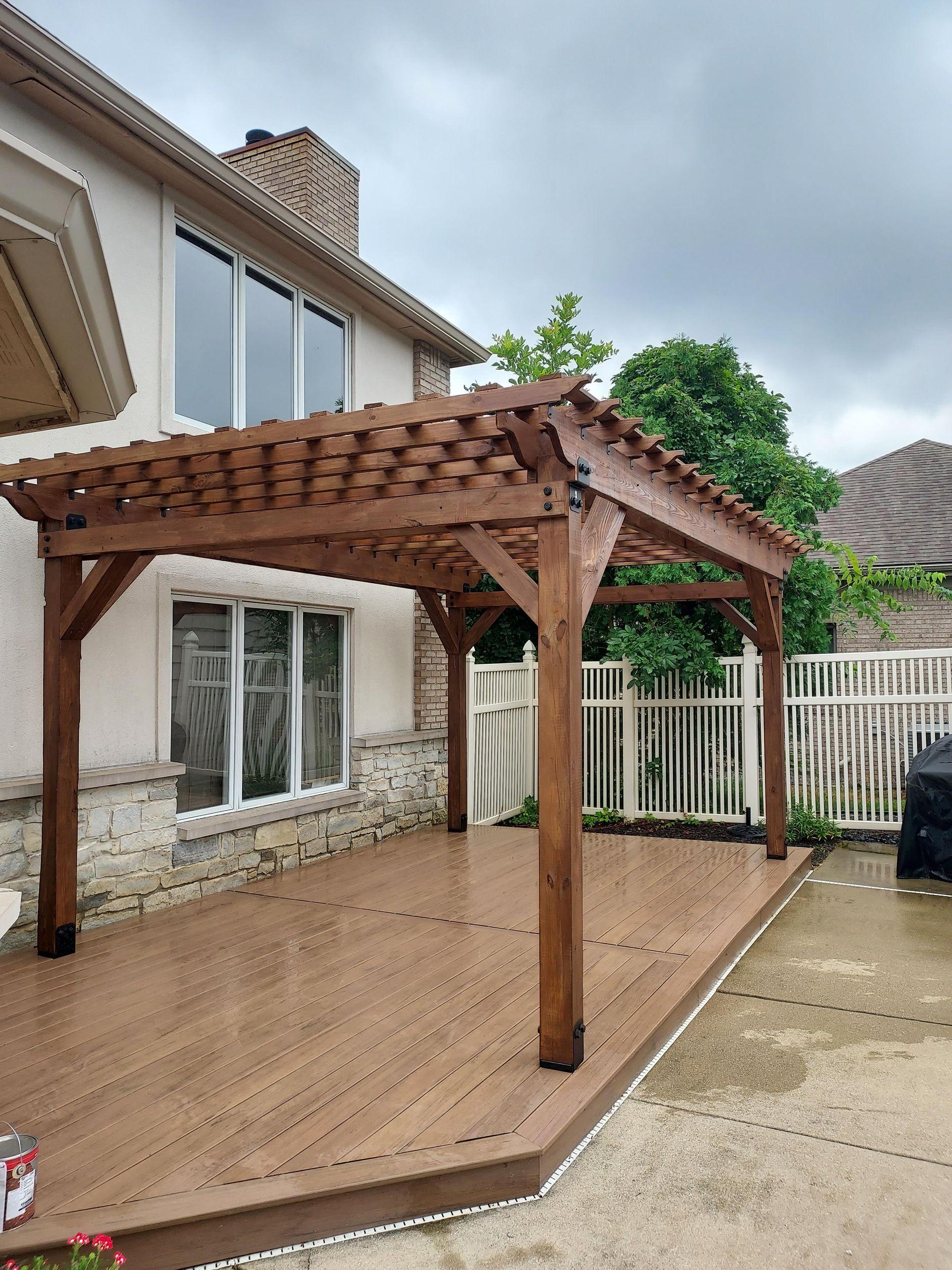 A wooden pergola covers a composite deck outside a two-story beige house. Overcast sky.