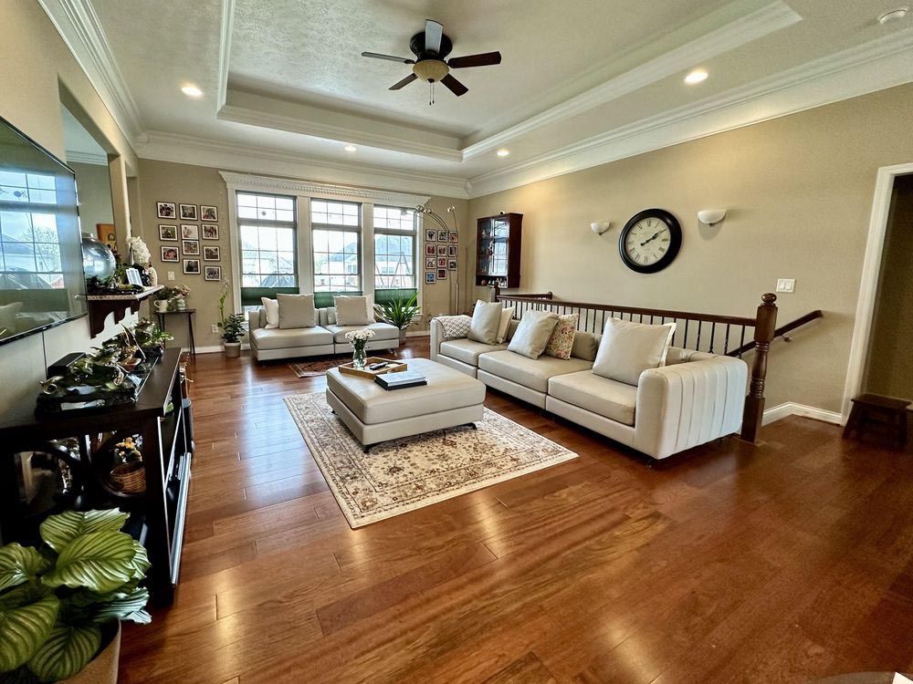 Living room with hardwood floors, beige sofas, and a rectangular ottoman on a patterned rug. A large clock hangs on the wall, and a staircase is visible.