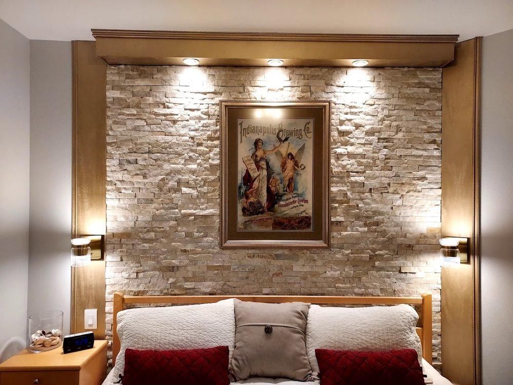 Bedroom wall with a stone accent, artwork, and decorative lighting above the bed with pillows and a bedside table.