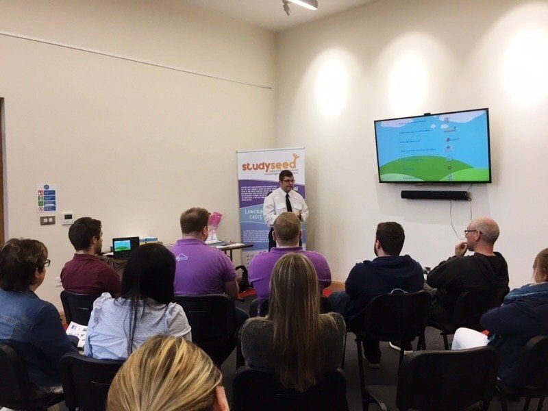 Man giving a presentation in a room with a TV screen and a small audience seated.