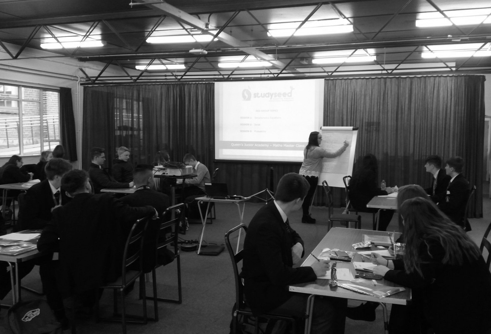 Students in suits attending a presentation in a classroom. A woman writes on a flip chart, projected content on screen.
