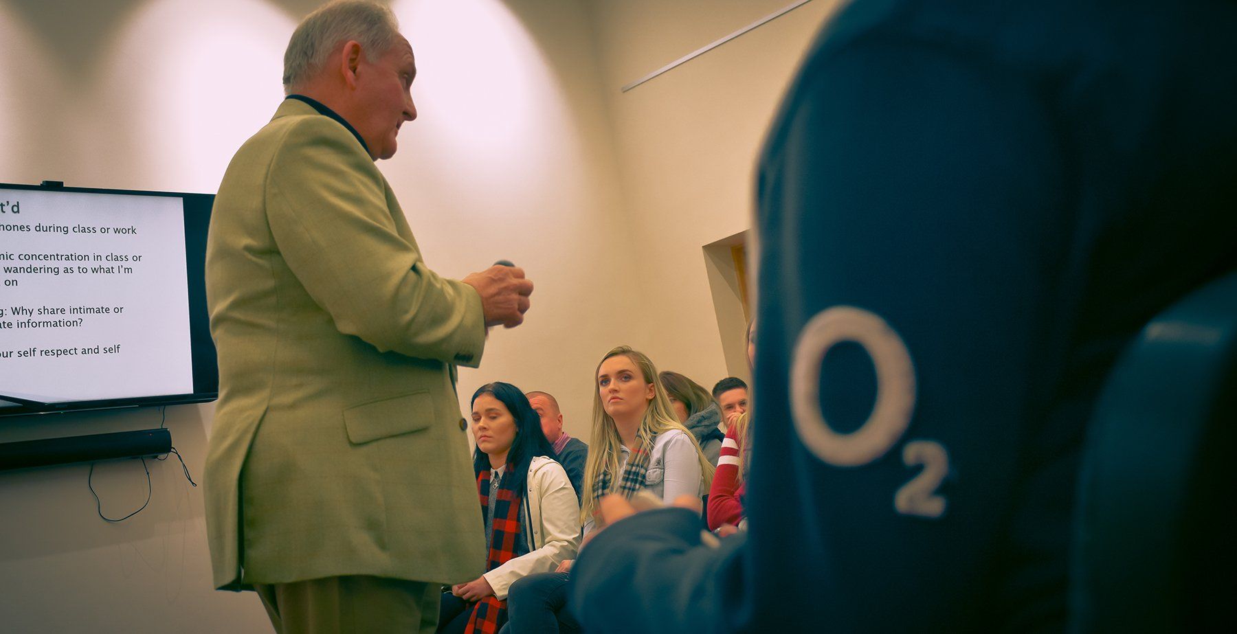 Man lecturing in front of a screen, facing seated audience. Person with O2 logo on sweatshirt.