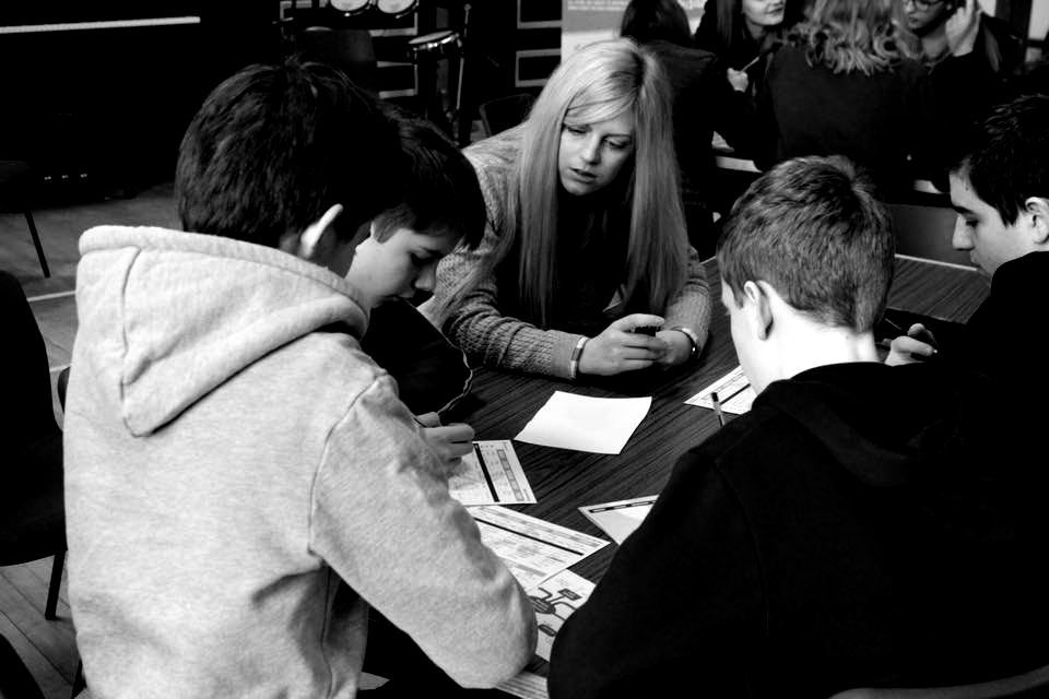Group of people at a table, looking at cards or papers. One person in a gray hoodie, others in dark clothing.
