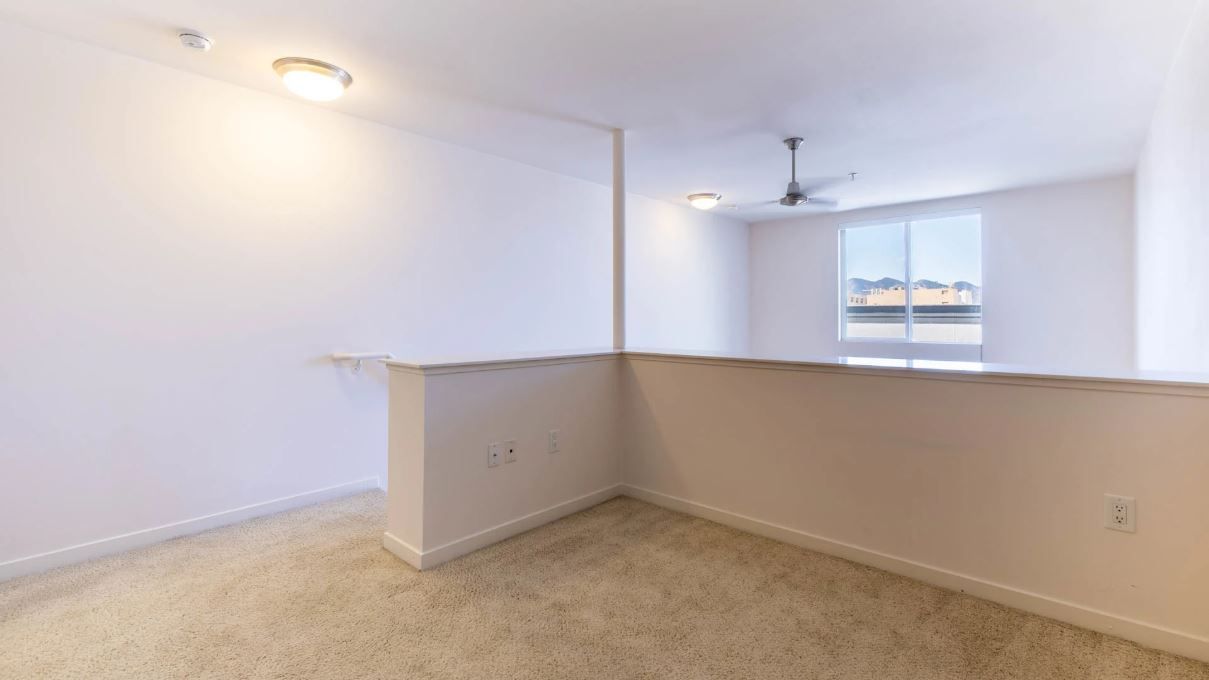 Empty loft area with white walls, tan carpet, and a window; a ceiling fan and lights are visible.