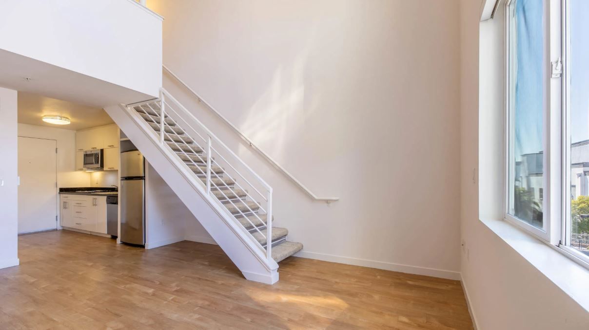 Loft interior: white staircase, kitchen, hardwood floors, large window with a cityscape view.