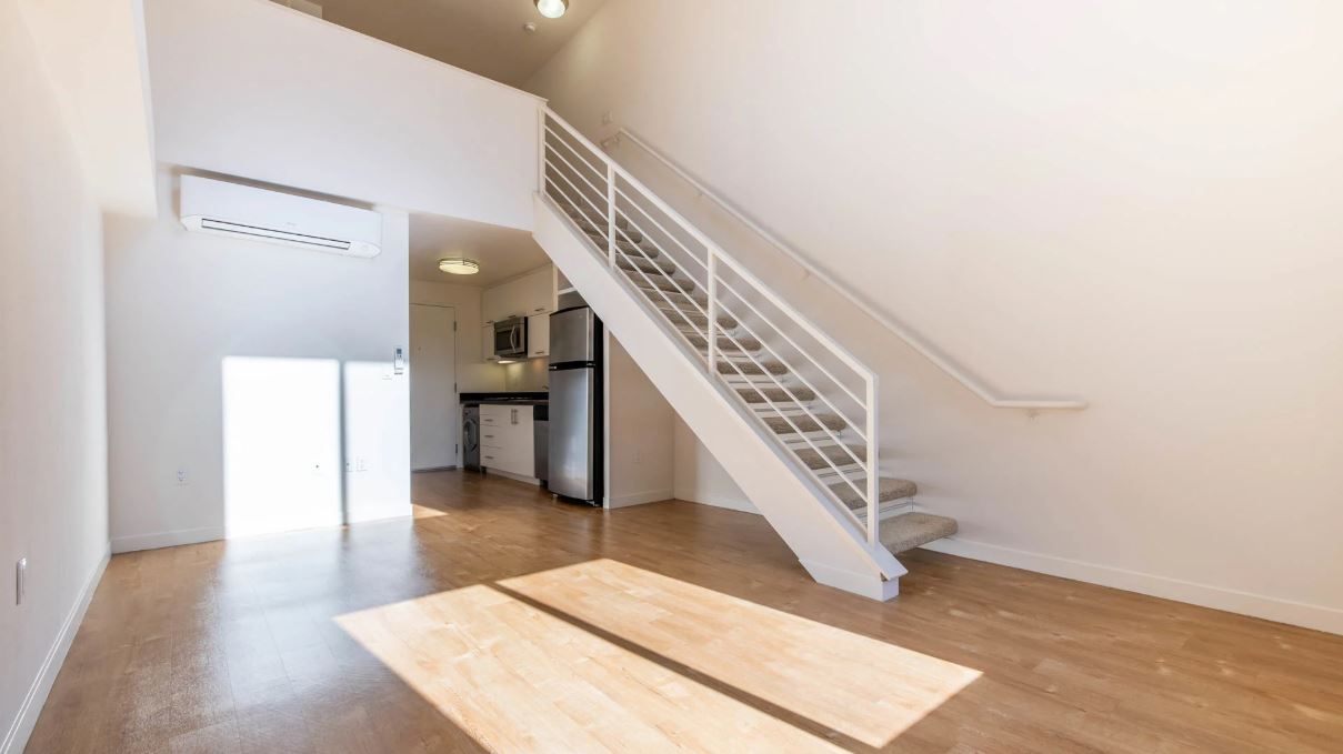 A two-story loft interior with white walls, wooden floors, stairs, and a small kitchen visible in the background.