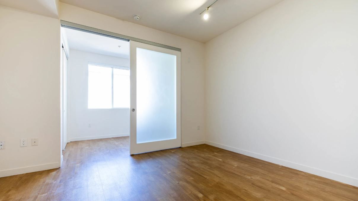 Empty room with hardwood floors and a frosted sliding door, adjacent to another room with a window.
