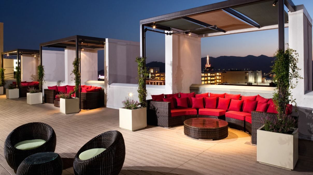Rooftop lounge with red couches, wicker chairs, and city views at dusk.