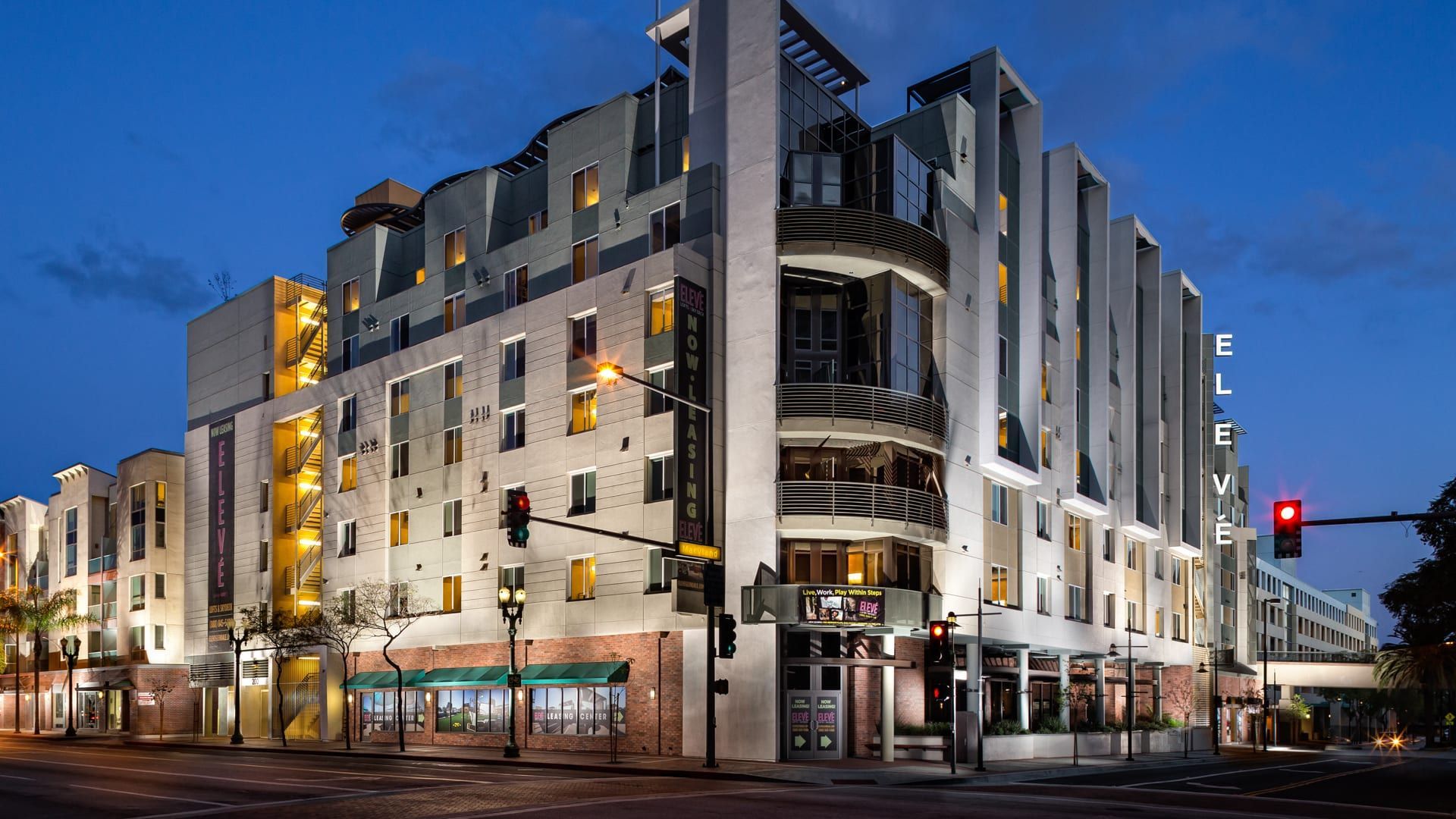 Apartment exterior at dusk with illuminated windows, traffic lights, and street lights.