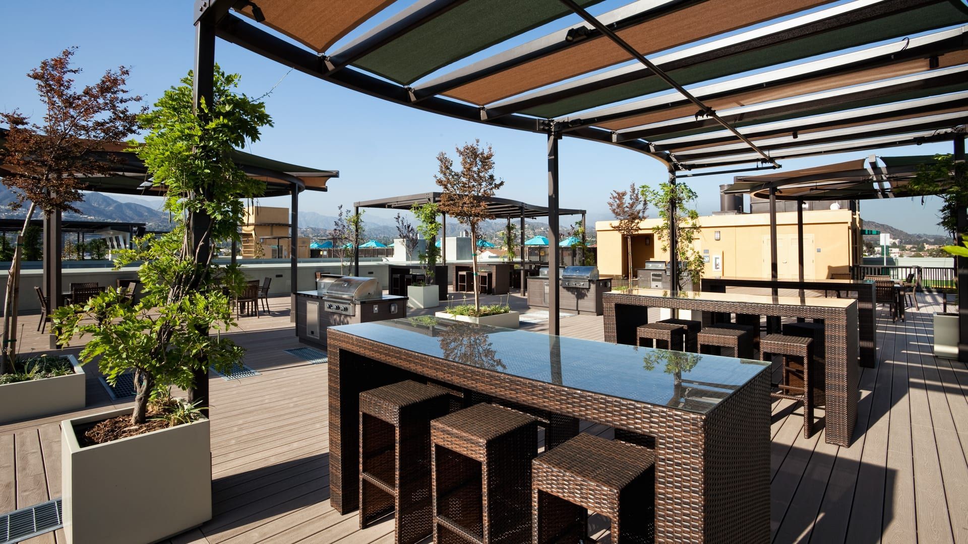 Rooftop patio with wicker tables and stools, shade structures, and potted trees. 