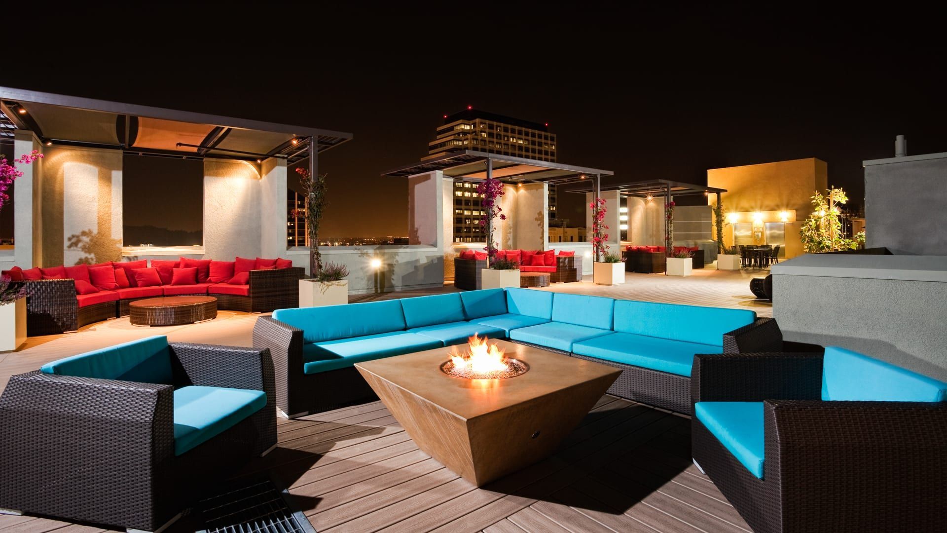 Rooftop lounge at night with turquoise and red couches, a fire pit, and city views.