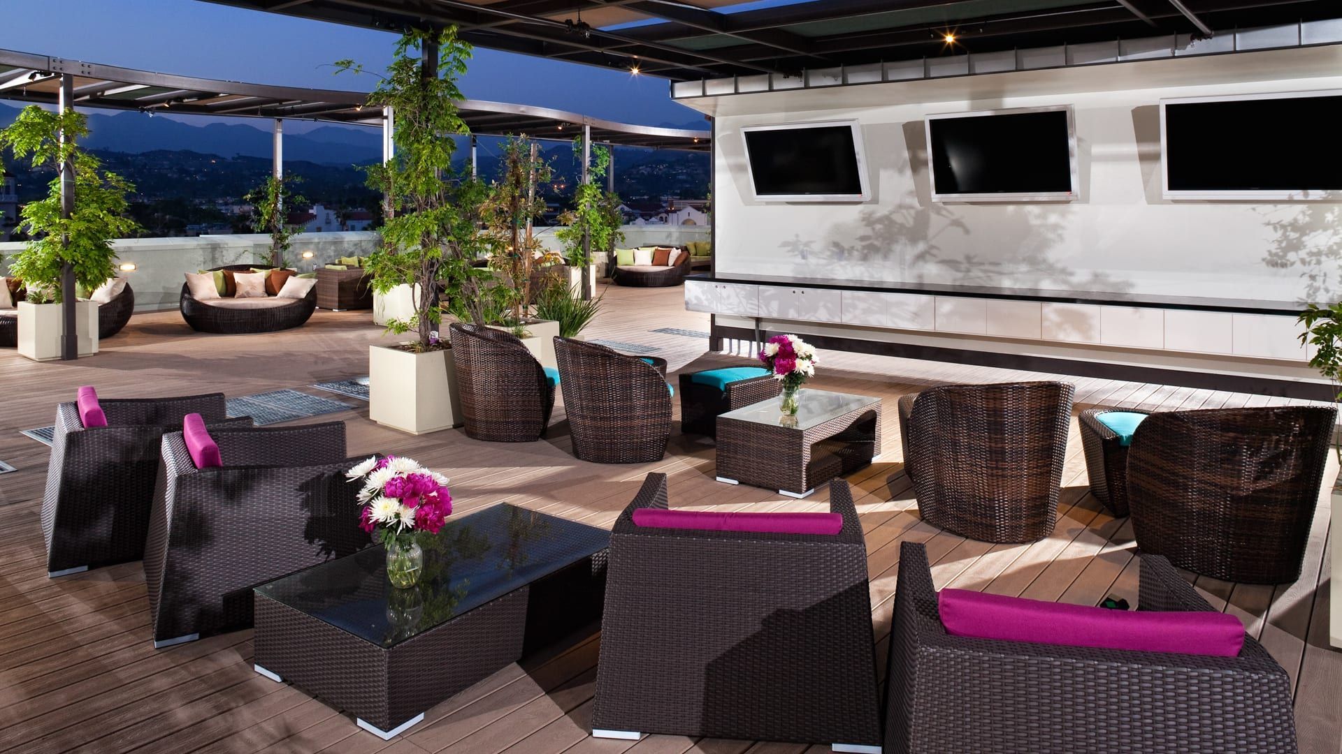 Rooftop lounge at dusk with wicker furniture, city views, and televisions.