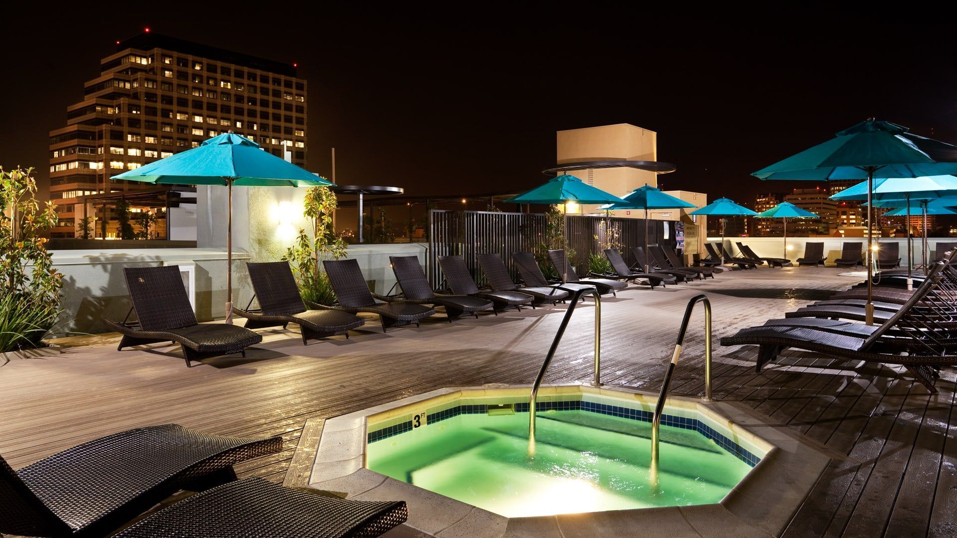 Rooftop pool and hot tub at night; turquoise umbrellas and city backdrop.