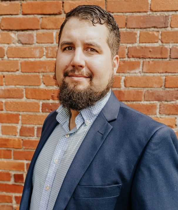 A person with a beard, wearing a blue blazer and light patterned shirt, posing in front of a brick wall.