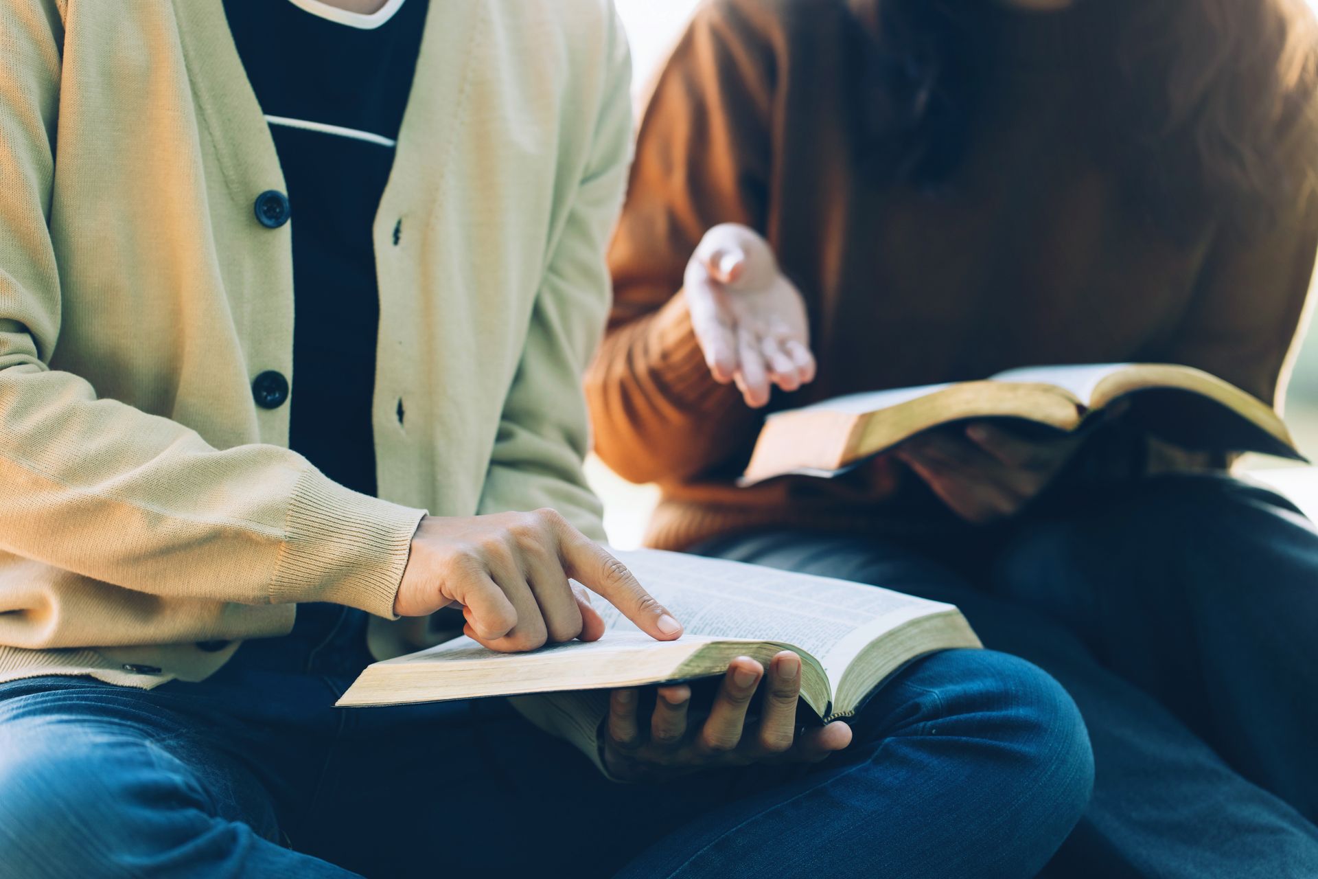 Two people seated outdoors, looking at open books. One points at a page, the other gestures with their hand.