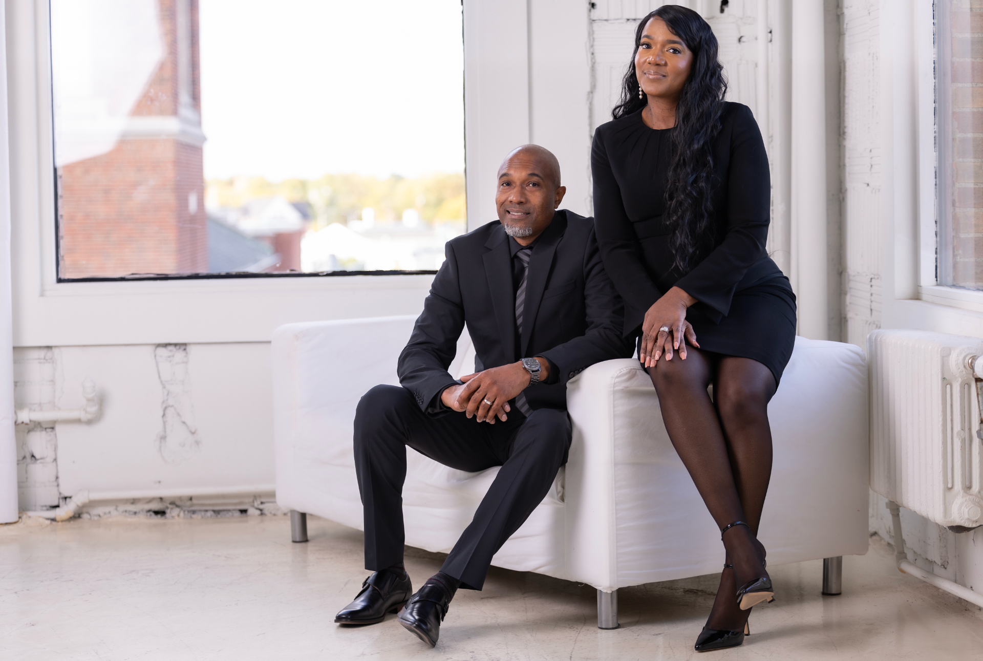 A man and woman sit on a white couch near a window.