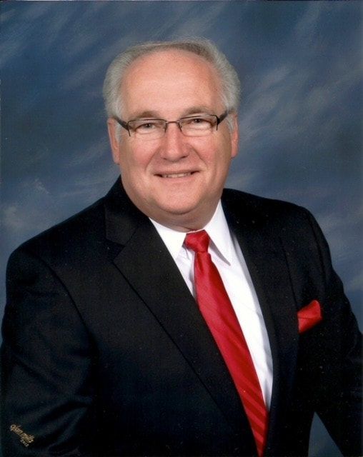 A man wearing a black suit, white shirt, red tie, and glasses poses against a blue mottled background.