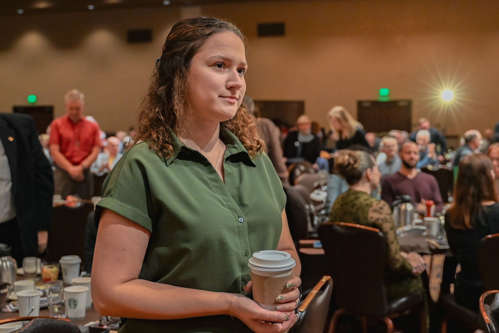 A woman in a green shirt is holding a cup of coffee in a conference room.