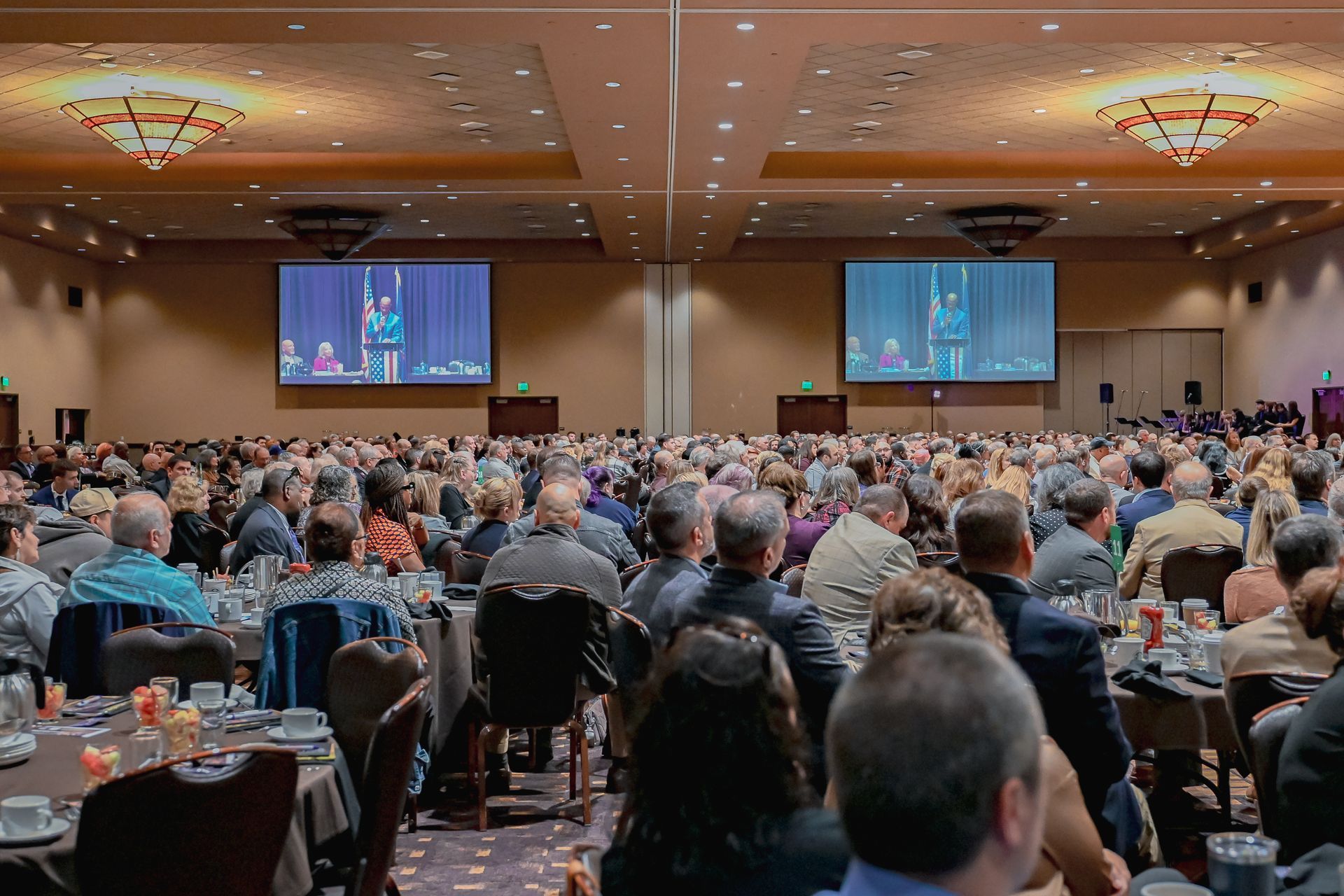 A large group of people are sitting at tables in a large room watching a presentation.