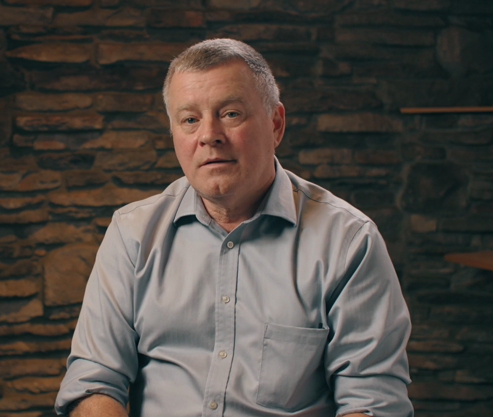 A man in a blue shirt is sitting in front of a stone wall.